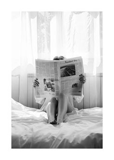 a woman sitting on top of a bed reading a newspaper with her legs crossed out