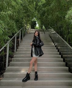 a woman in black jacket and skirt standing on stairs with trees behind her at the bottom