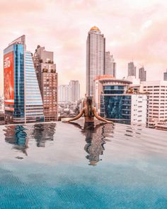 a woman is sitting on the edge of a swimming pool in front of tall buildings