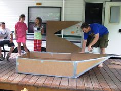 a group of people standing around a cardboard boat on a wooden deck with a trailer in the background