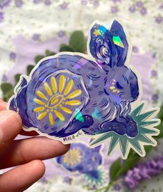 a hand holding a purple and yellow sticker in front of some flowers on a table