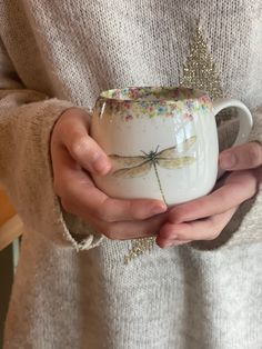 a woman holding a coffee cup with a dragonfly on it's side, in her hands