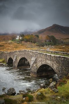 an old stone bridge over a river in the middle of a field with mountains behind it