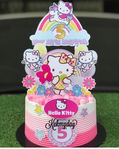 a hello kitty birthday cake on top of a green field with flowers and rainbows