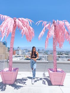 a woman standing next to two pink palm trees on top of a roof with buildings in the background