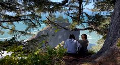 two people sitting on the side of a mountain looking out at the ocean and trees