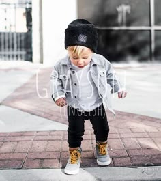 a little boy standing on top of a sidewalk wearing a black hat and jacket with yellow laces