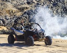 two people in a can - am off road vehicle driving on the sand near some rocks