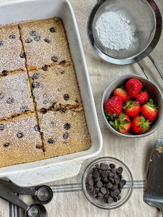 a pan filled with cake next to bowls of strawberries and chocolate chips