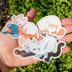 a hand holding three stickers with cats and kittens on them in front of some bushes