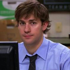 a man wearing a blue shirt and tie sitting in front of a computer monitor looking at the camera