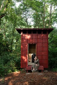 a person sitting on a bench in front of a red outhouse surrounded by trees