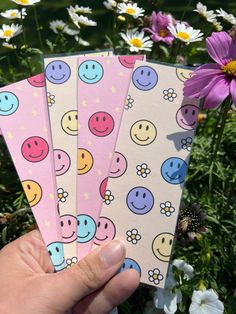 three notebooks with smiley faces on them are held in front of some daisies