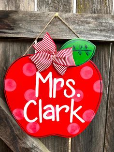 a red apple shaped door hanger with the words mrs clark painted on it and a bow