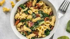 a white bowl filled with pasta and spinach next to a fork on a table