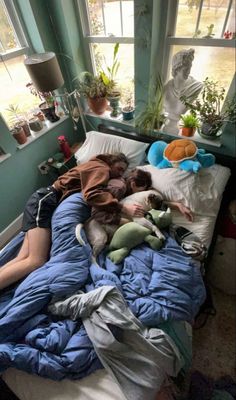 two people sleeping on a bed with stuffed animals and plants in the window sill