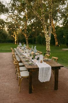 a long wooden table with white linens and flowers on it is surrounded by string lights