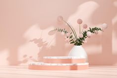 a white vase with pink flowers in it sitting on a table next to a wall