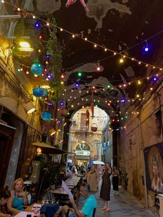 people are sitting at tables in an alleyway with lights strung from the ceiling above them