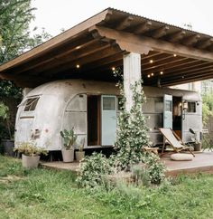 an old camper sits in the grass under a covered area with lounge chairs and potted plants