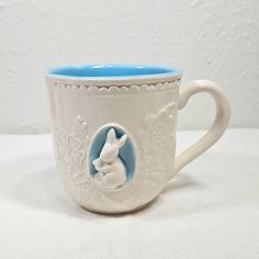 a white and blue coffee cup with a rabbit on it's side, sitting on a table