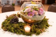 a glass bowl filled with flowers and greenery on top of a table next to a candle