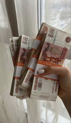 a person holding up five stacks of money in front of a window with white curtains