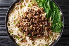 a bowl filled with noodles and meat on top of a wooden table next to spinach leaves