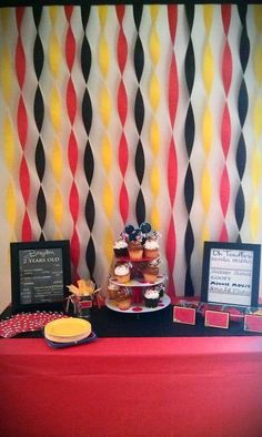 a red table topped with lots of cupcakes next to a wall covered in black and yellow stripes