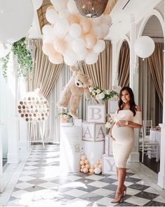 a woman in a white dress standing next to balloons and a teddy bear on the wall