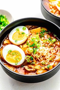 two bowls filled with ramen and eggs on top of a white table next to green onions