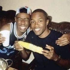 two men are sitting on a bed and smiling at the camera, one is holding a corn cob