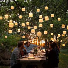 people sitting at a table with paper lanterns hanging from the trees in the evening time