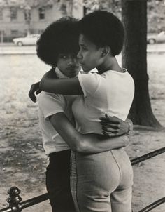 two young men embracing each other in front of a tree and fence with cars parked on the street behind them