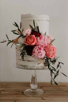 a white cake with pink flowers on top and greenery around the edges, sitting on a wooden table