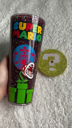 a hand holding a tumbler cup with the words super mario printed on it and a button