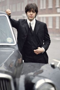 a man in a suit and tie leaning on the hood of a car with his hand up