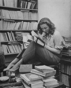 a woman sitting on the floor reading a book and talking on her cell phone in front of a bookshelf