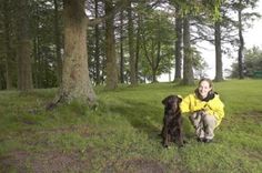 a woman kneeling down next to a dog in the grass with trees behind her on a sunny day