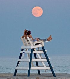 two people are sitting in chairs on the beach under a full moon, with their backs to each other