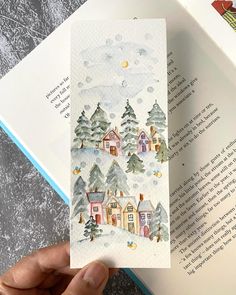 a hand holding an open book with watercolor houses and trees on the pages in front of it