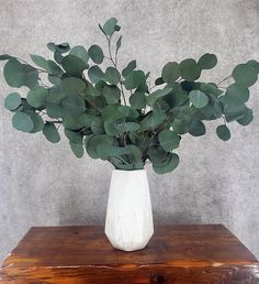 a white vase filled with green leaves on top of a wooden table