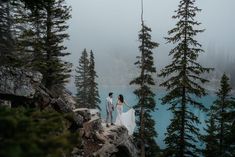 a bride and groom standing on the edge of a cliff overlooking a lake surrounded by trees