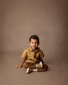 a little boy sitting on the ground with his legs crossed and smiling at the camera