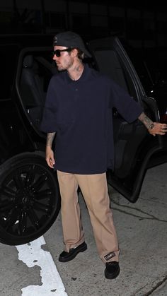 a man in black shirt and tan pants standing next to a car with his hand on the door