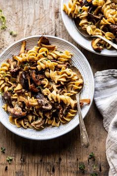 two white plates filled with pasta and mushrooms on top of a wooden table next to silverware