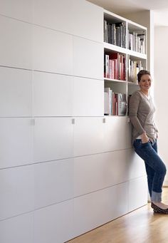 a woman leaning against a book shelf in a room with white walls and wooden floors