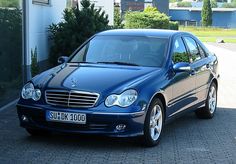 a blue mercedes benz parked in front of a building with trees and bushes behind it