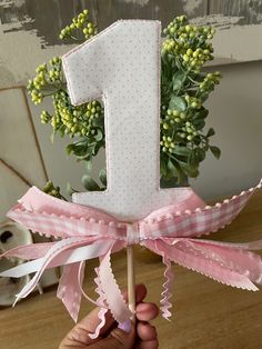 a hand holding a pink and white number one cake topper with flowers in the background