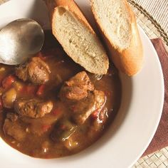 a white plate topped with meat and vegetable soup next to a piece of bread on top of a table